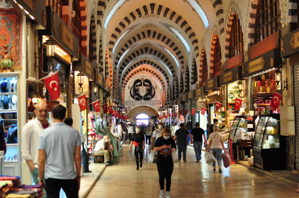 A crowded scene inside the main corridor of the Spice Bazaar, where tourists and locals walk past colorful shops decorated with Turkish flags and an Ottoman-era talismanic symbol hanging in the background. photo by Serhat Engul