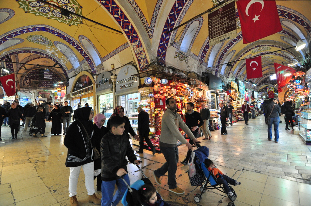 A crowded tourist family walking through one of the intersecting streets inside the Grand Bazaar while curiously observing the surrounding shops, with the yellow and dark blue ceiling decorations visible before the restoration repainting of the early 2020s. photo by Serhat Engul