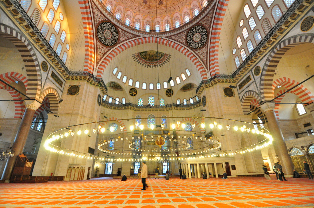 Wide-angle interior view of Süleymaniye Mosque looking toward the mihrab, with the massive chandelier hanging low above the prayer hall while a passing visitor provides a sense of the building’s monumental scale, photo by Serhat Engul