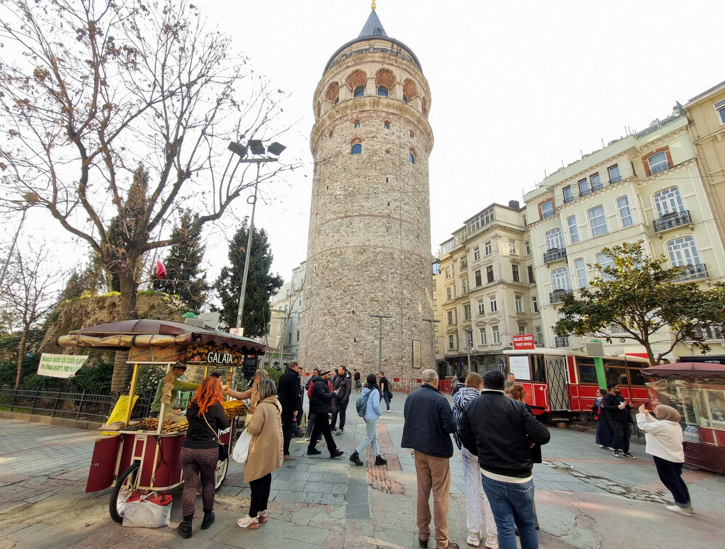 Tourists shopping from a street vendor and buying tickets from the nostalgic tram-shaped ticket booth in the lively Kuledibi Square beneath Galata Tower. photo by Serhat Engul