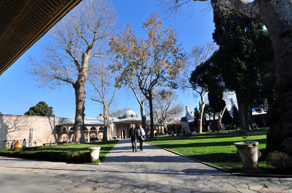 A couple walking along the stone pathway in the Second Courtyard of Topkapı Palace with the Gate of Felicity (Babüssaâde), which leads to the Third Courtyard, visible behind them, photo by Serhat Engul
