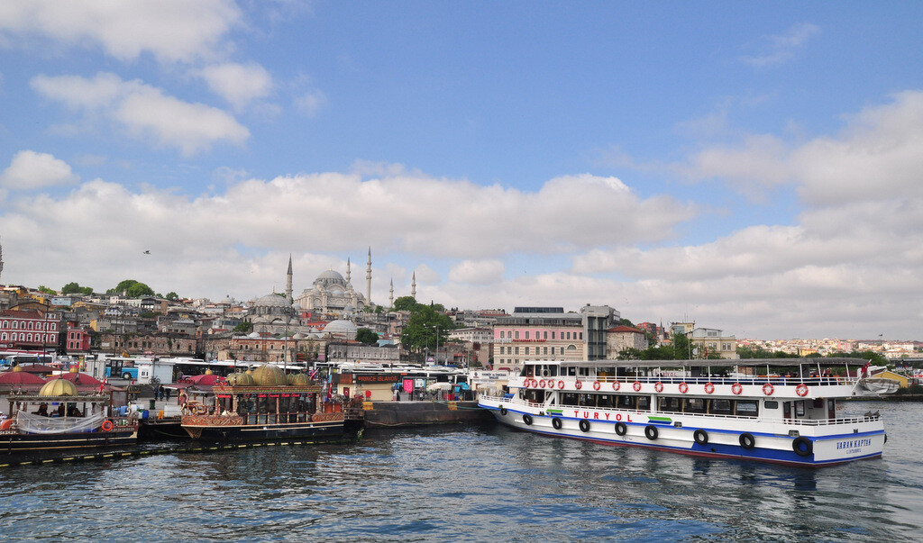 Turyol Bosphorus cruise boat docked at the Eminönü pier beside traditional fish sandwich boats, with Süleymaniye Mosque rising above the Historical Peninsula in the background, photo by Serhat Engul
