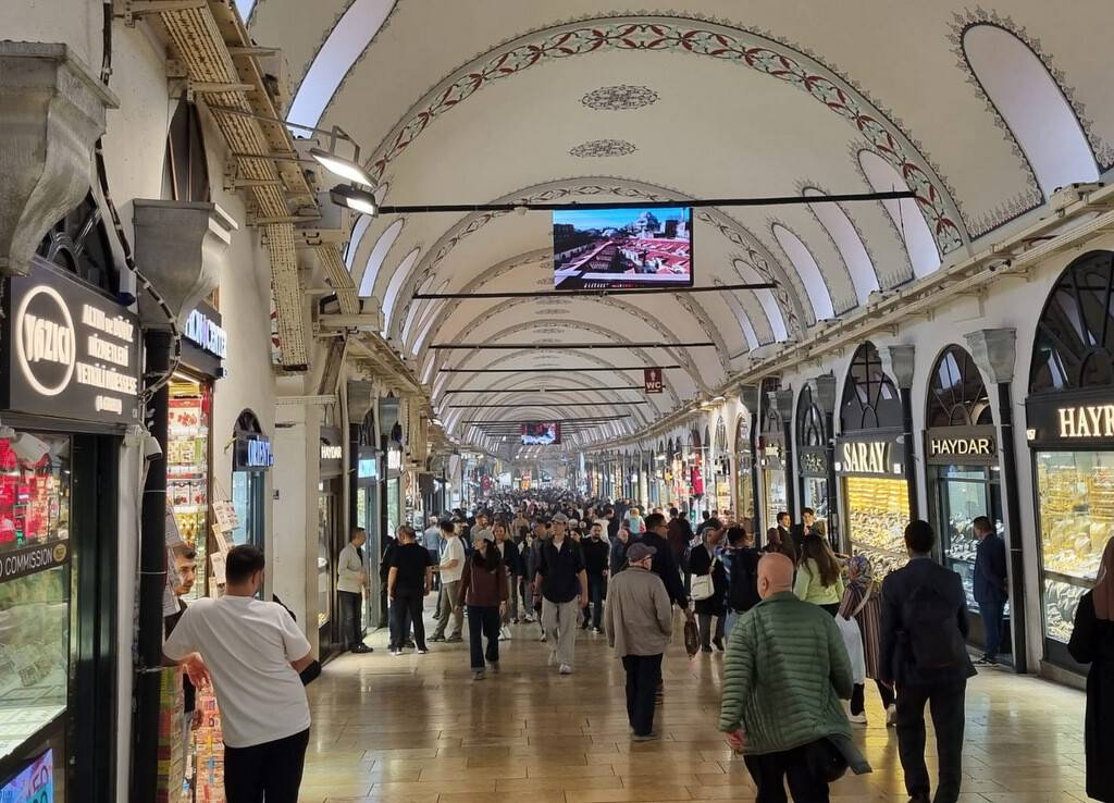 Tourists walking downhill along Kalpakçılarbaşı Street inside the Grand Bazaar after entering from the Beyazıt side through Gate 7, surrounded by colorful shops on the way toward the Nuruosmaniye Gate. photo by Serhat Engul
